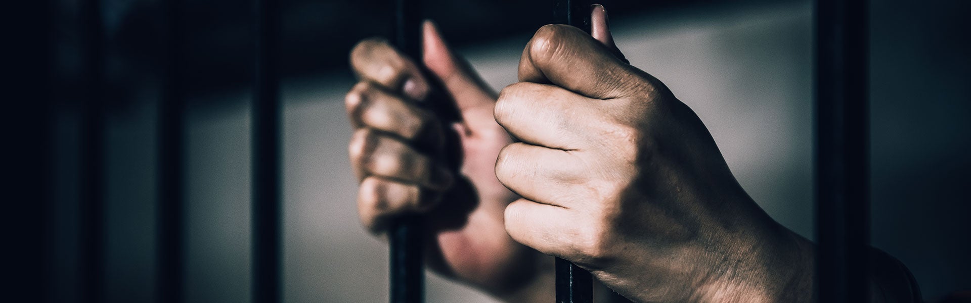 A person's hand clutches the bars of a dark metal jail cell.