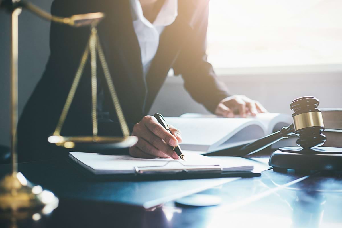 A person in a suit writes on paperwork with a pen next to a gavel and scales of justice.