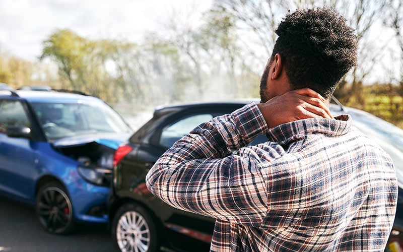 A man with a concerned expression massages his neck near two damaged cars at an accident scene.