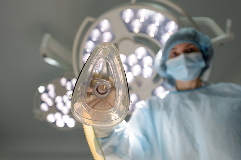 A medical professional wearing a surgical mask and cap holds an oxygen mask in an operating room.