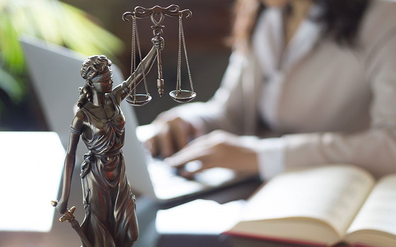 Lady Justice statue with scales balances over a blurred background of a person typing on a laptop.