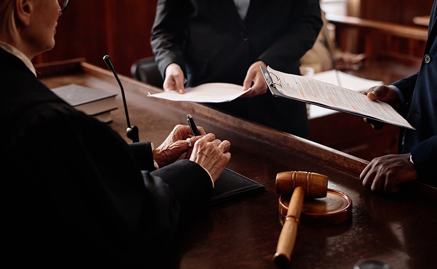 A woman in a suit takes notes at a table while a man presents papers, a gavel rests nearby.