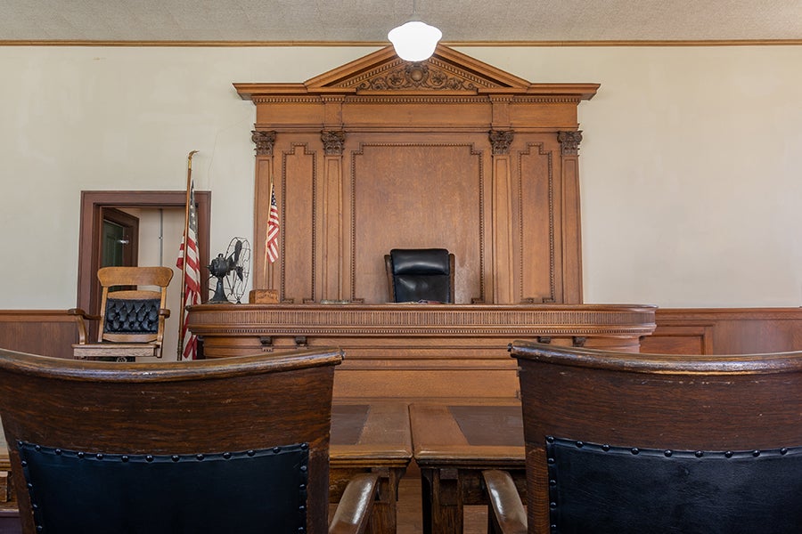 A wooden judge's bench sits centered in a courtroom, with rows of chairs visible in the foreground.
