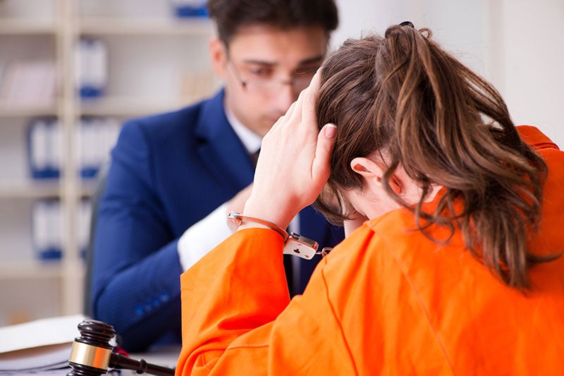 A woman in an orange jumpsuit touches her forehead while speaking with a man in a suit across a desk.