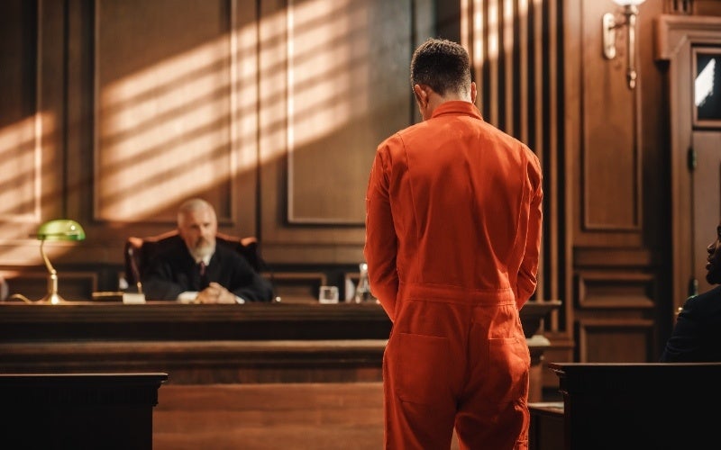 A person in an orange jumpsuit stands facing away, seemingly in a courtroom with a judge and another person present.