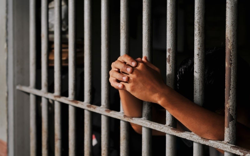 A person with dark skin clasps their hands together behind metal prison bars.
