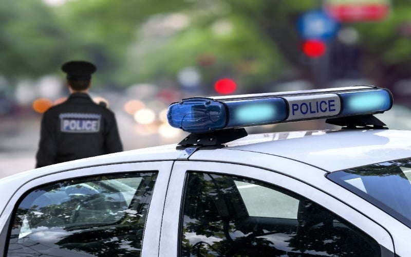 A police car’s flashing blue and white lights are visible with a police officer standing in the background.