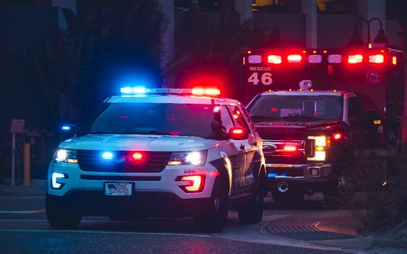 An emergency vehicle with flashing red and blue lights sits parked next to a police car at night.