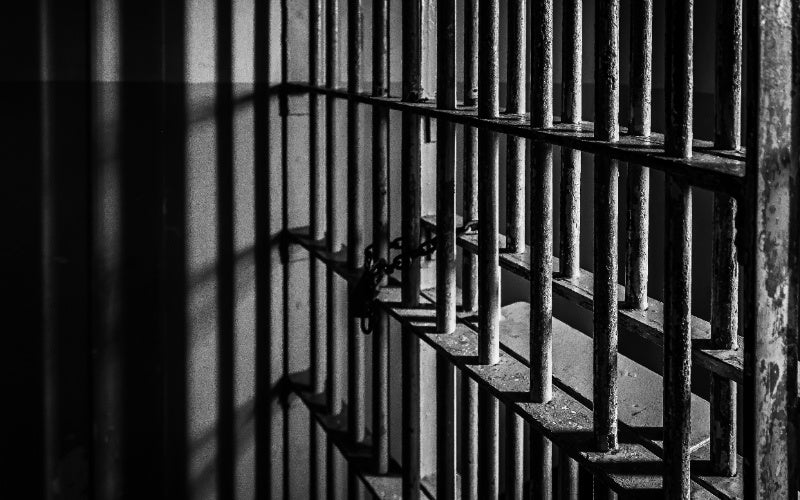 Shadows fall across a dark prison cell with metal bars and a heavy chain lock.