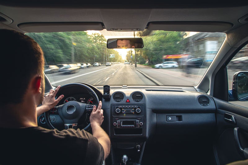 A person drives a car down a tree-lined road with sunlight streaming ahead.