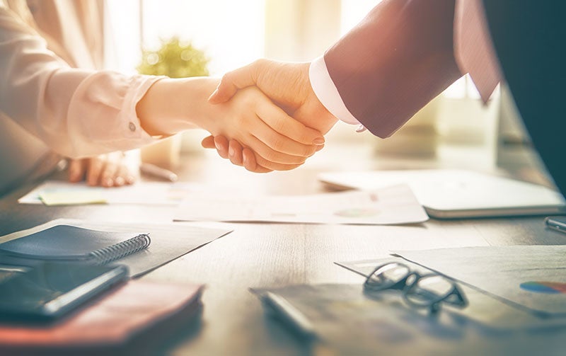 Two people shake hands across a table covered with documents and office supplies.