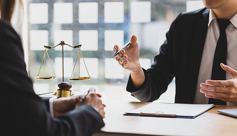 A lawyer in a suit gestures while speaking to a client across a table with legal documents and scales of justice in the background.