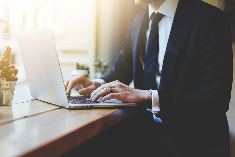 A man in a suit types on a laptop while seated at a wooden table.