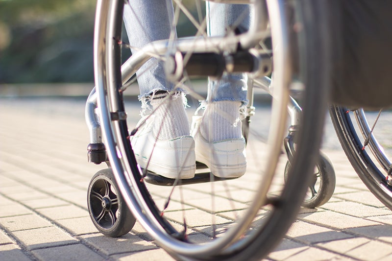 Legs in white sneakers rest on a wheelchair footrest on a paved walkway.
