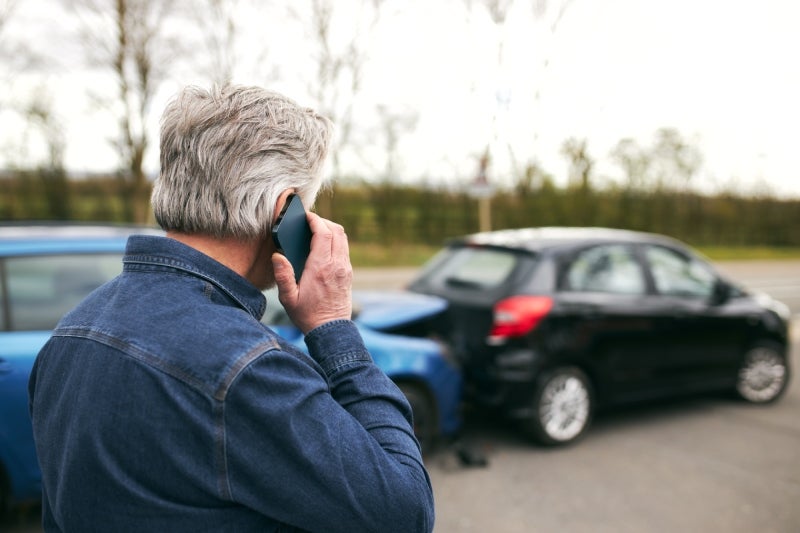 An older man with gray hair speaks on a mobile phone while standing near a blue car and a damaged black car.