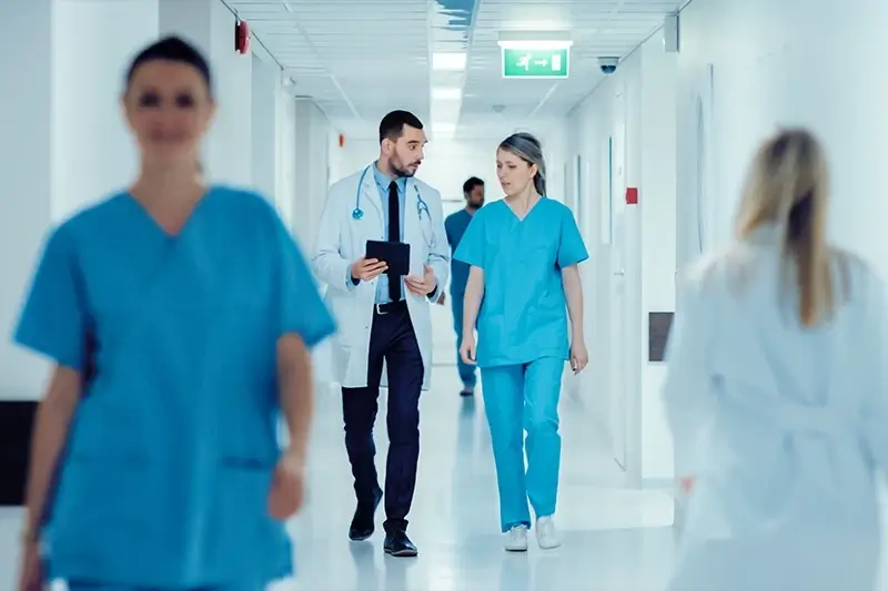 A doctor and nurse walk down a brightly lit hospital hallway, discussing a tablet.