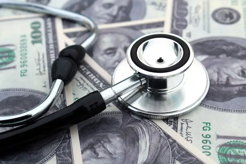 A silver stethoscope rests on a background of hundred-dollar bills.