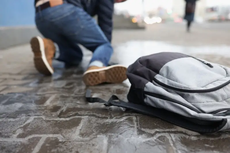 A person kneels on a cobblestone street next to a gray backpack that has fallen to the ground.