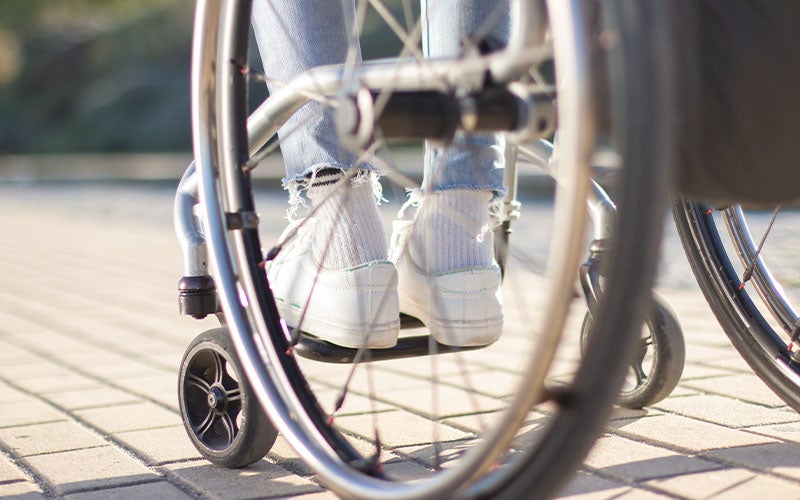 Feet in white sneakers rest on a wheelchair footrest on a paved path.