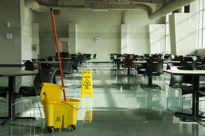 A yellow wet floor sign sits near a mop and bucket in a large, empty cafeteria.