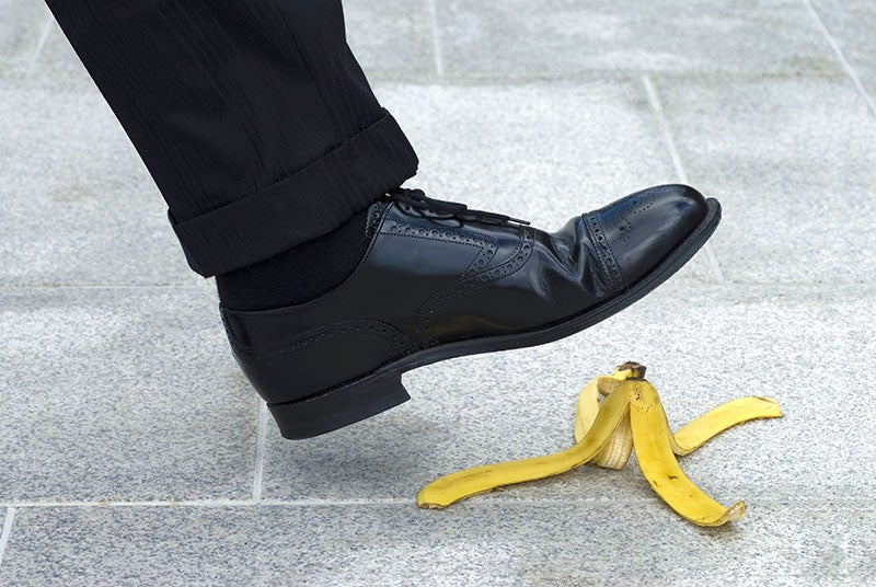 A person in black dress shoes appears to be stepping on a discarded banana peel on a tiled surface.